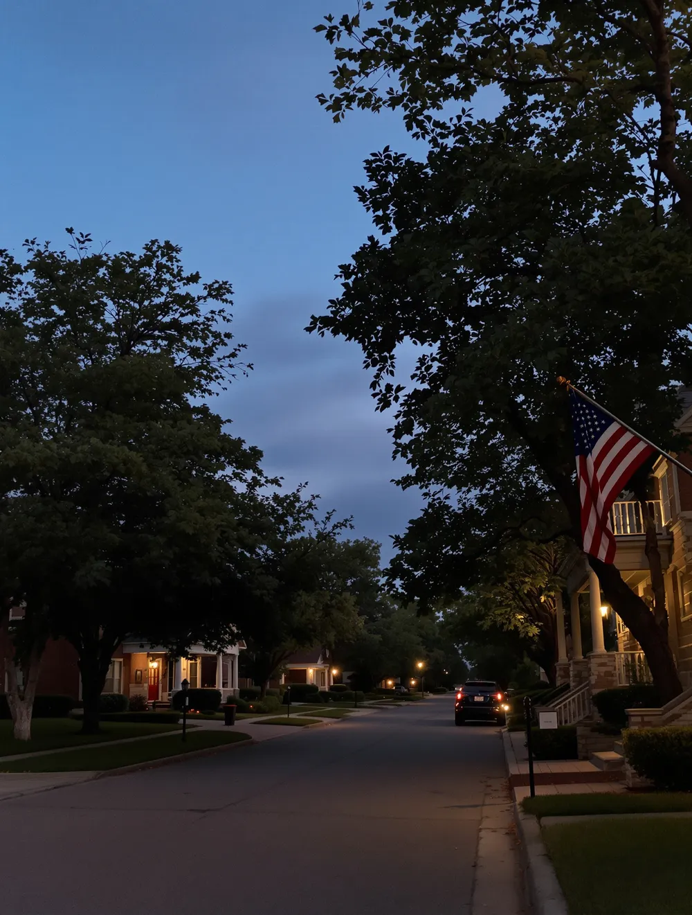 A quiet established North Texas residential street at dusk, brick and stone homes with mature trees and an American flag mounted by a front door, warm porch light beginning to glow.