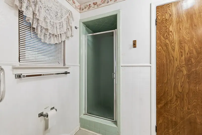 Tiled stall shower with a glass door beside a lace-curtained window