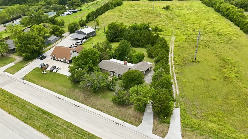 Wide aerial of the home, driveway and open green field with neighboring buildings along the highway