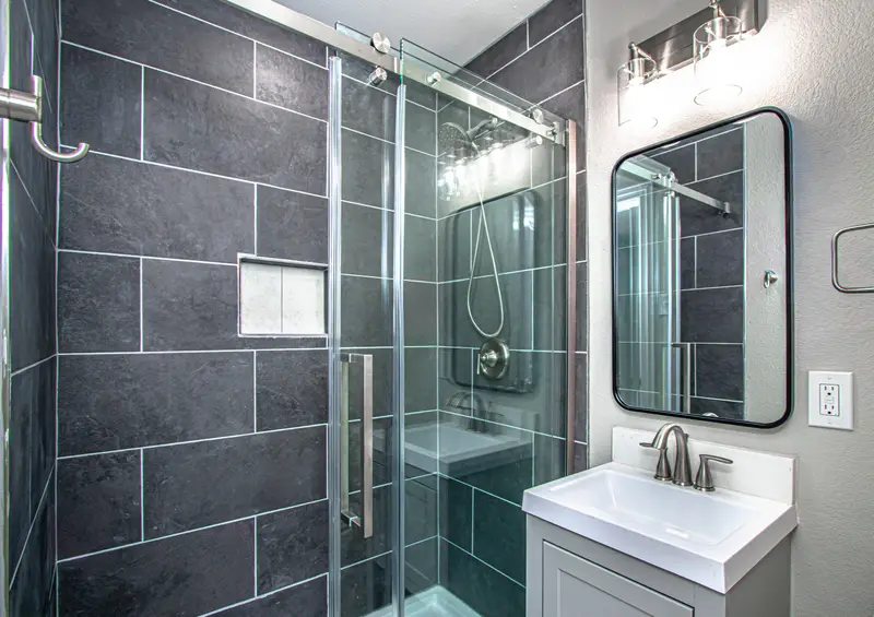 Glass walk-in shower with dark large-format tile and a niche, beside a gray vanity and black-framed mirror