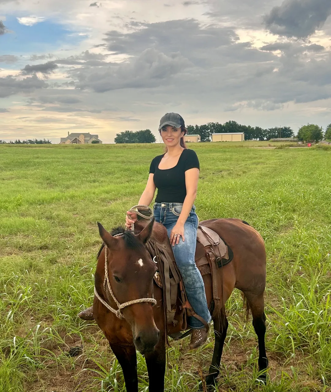 Nychole Baxter on horseback in a green Texas pasture at dusk, a ranch house on the horizon behind her.